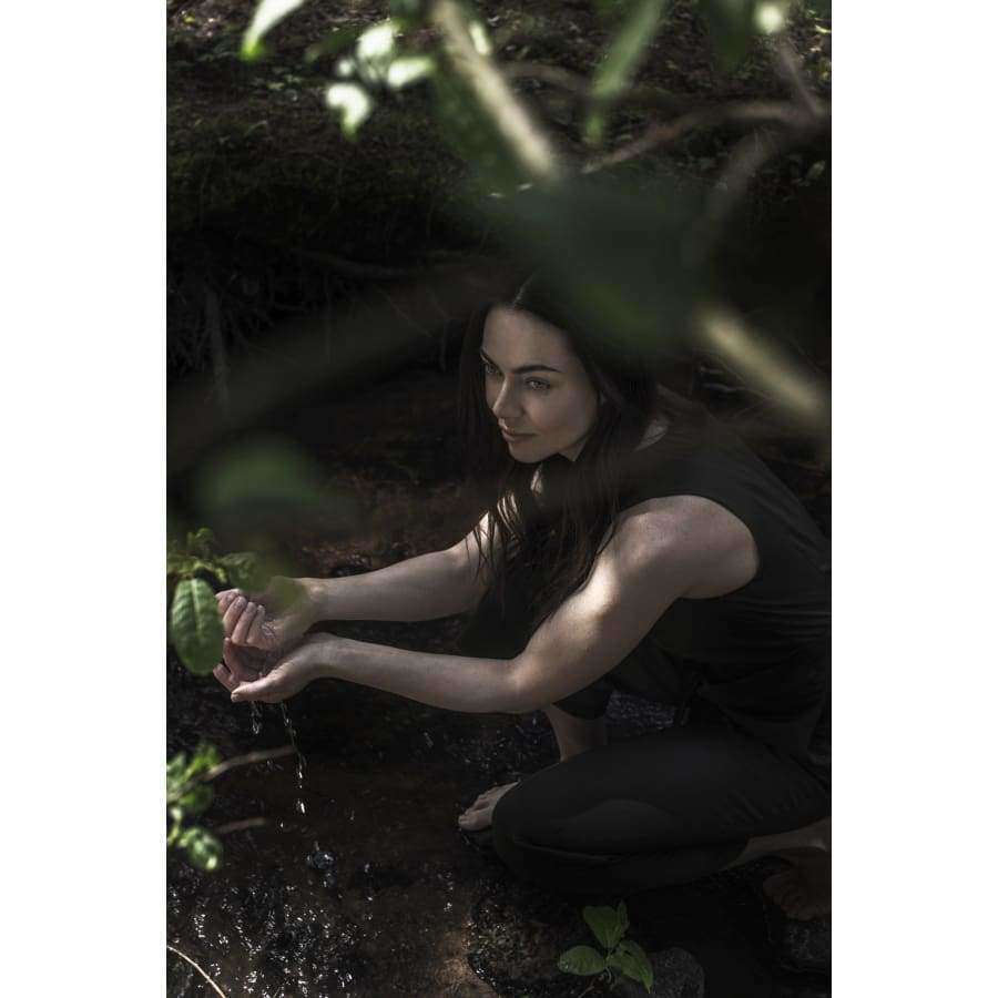Een vrouw hurkt bij een beekje in het bos en schept water op met haar handen terwijl het zonlicht door de bladeren boven haar hoofd schijnt. Omringd door groen denkt ze aan de verfrissende zuiverheid van Taika Liquid Hand Soap van Taika.